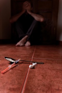 Man lying on the floor in the dark with his hands on his head desperate for drugs in the foreground Syringe with heroin and spoon