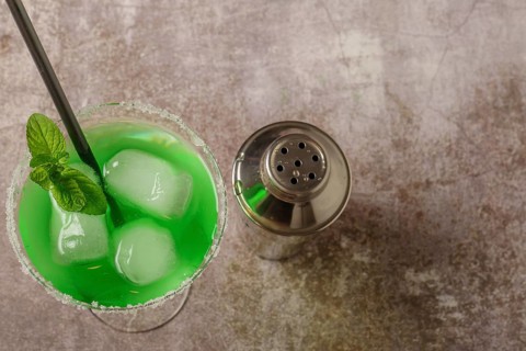 Top view of a green cocktail with mint ice and straw the edges of the glass have icing sugar and a cocktail shaker to accompany it
