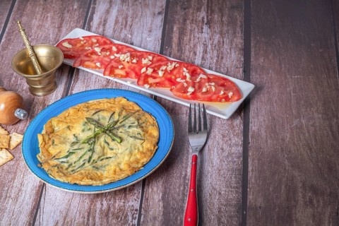 Wild asparagus omelette on a blue plate with a tomato salad with garlic on a wooden table