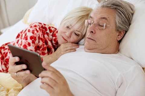 Elderly couple lying together in bed in bedroom looking at smartphone together