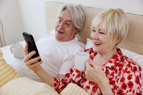 Elderly couple lying together in bed in bedroom looking at smartphone together