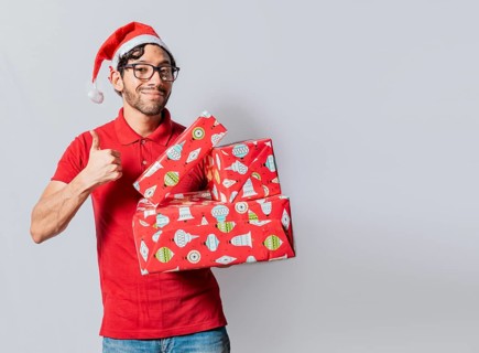 Christmas man holding gift boxes and smiling with thumb up. Smiling guy in christmas hat holding christmas gift boxes giving thumbs up. Friendly man in christmas clothes holding gift boxes