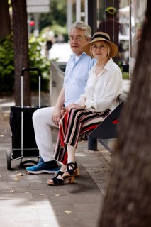 Older couple dressed in summer clothes with a trolley waiting for a bus at a bus stop