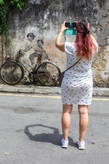 Young woman taking a photo of street art boy and girl on bicycle on a wall in George Town on Penang Island