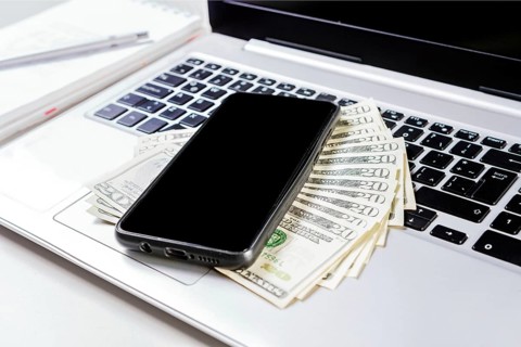 Cell phone on laptop keyboard and dollar bills. Mobile phone on top of dollar bills on laptop keyboard. Top view of laptop with cell phone and dollar bills