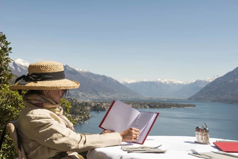 Woman with Straw Hat Sitting and Reading the Menu in a Restaurant with Panoramic View over Lake Maggiore in a Sunny Day in Ascona