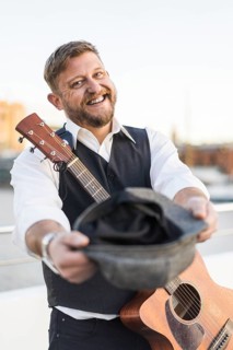 A smiling street musician looking at camera and carrying his guitar while asking for tips