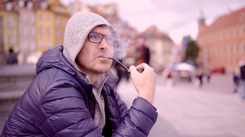 Adult man with glasses sitting on square and smoking a tobacco pipe releasing smoke in the Palace Square