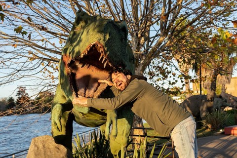 Latin man at an amusement park making himself enter the mouth of a fake dinosaur
