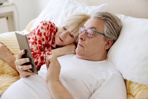 Elderly couple lying together in bed in bedroom looking at smartphone together