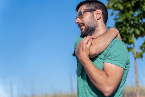 Profile view of an attractive bearded man with sunglasses stretching his shoulders in a field with blue sky in the background