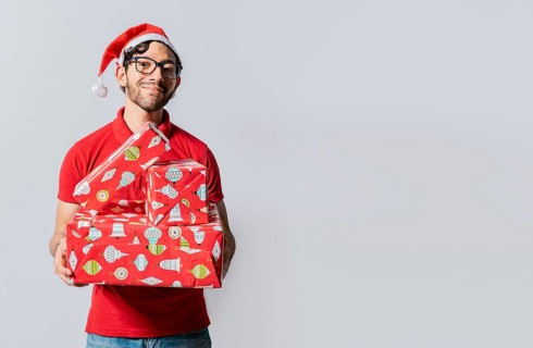 Christmas man portrait holding gift boxes and smiling isolated. Smiling guy in santa hat holding christmas gift boxes. Friendly man in christmas clothes holding gift boxes isolated