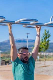 Bearded man with sunglasses seen from the front training his back by doing pull-ups on a barbell assisted by an elastic fitness band in an outdoor gym