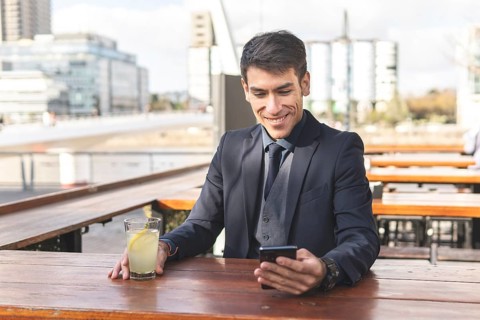 Businessman sitting at an outdoor bar drinking lemonade looking at his phone smiling