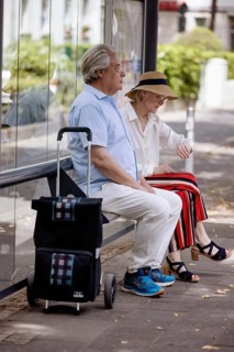 Older couple dressed in summer clothes with a trolley waiting for a bus at a bus stop