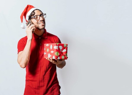 Smiling guy in christmas hat holding christmas gift box and talking on the phone. Christmas man holding gift and calling family on phone. Happy man holding gift and calling family at christmas