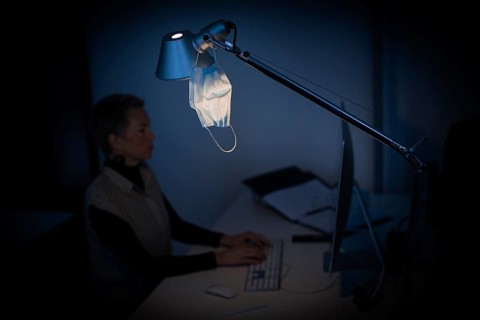 Mask at the workplace. A mask hangs from a lamp above a desk while a woman works on a computer in the background. Berlin