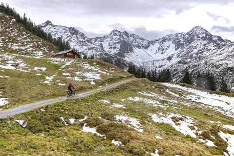 Two mountain bikers riding on path past wooden cabin on mountain slope with patches of melted snow in Montafon region in autumn