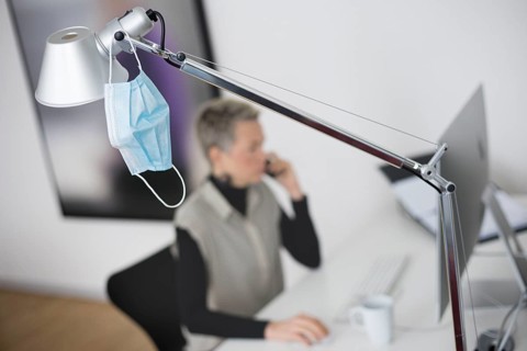 Mask at the workplace. A mask hangs from a lamp above a desk while a woman works on a computer in the background. Berlin