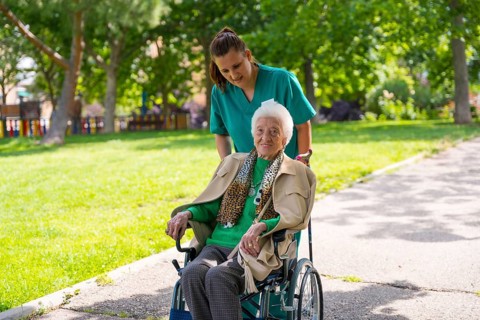 An elderly woman with the nurse on a walk through the garden of a nursing home in a wheelchair next to nature and trees