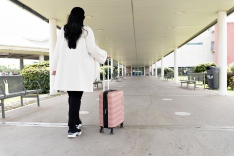 Back shot of woman in sneakers and coat walking towards main entrance of airport and rolling her wheeled suitcase. Travel concept