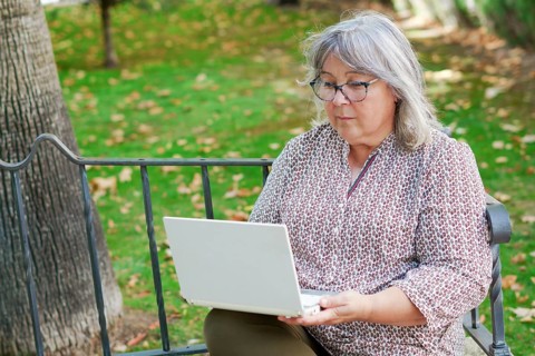 Older white-haired woman working on her laptop computer seated on a park bench