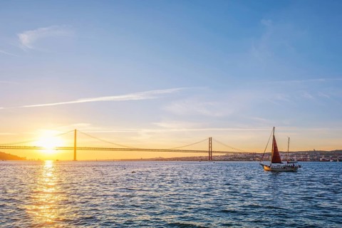 View of 25 de Abril Bridge famous tourist landmark of Lisbon connecting Lisboa and Almada over Tagus river with tourist yacht silhouette at sunset. Lisbon