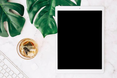 Brewing hot beverage with tea bag glass monstera leaves keyboard digital tablet white desk