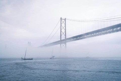 View of 25 de Abril Bridge famous tourist landmark of Lisbon connecting Lisboa and Almada in heavy fog mist wtih yacht boats passing under. Lisbon