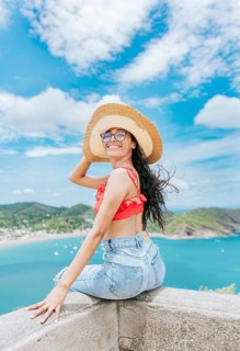 Portrait of tourist girl in a hat at a viewpoint with a beach in the background. Happy tourist female sitting at a viewpoint with beach in the background