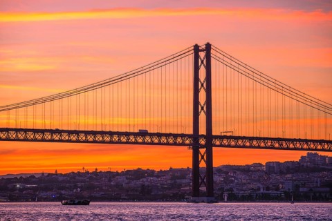View of 25 de Abril Bridge famous tourist landmark of Lisbon connecting Lisboa and Almada over Tagus river with tourist boat silhouette at sunset. Lisbon
