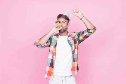 Man raising his arms while blowing party horn pink background