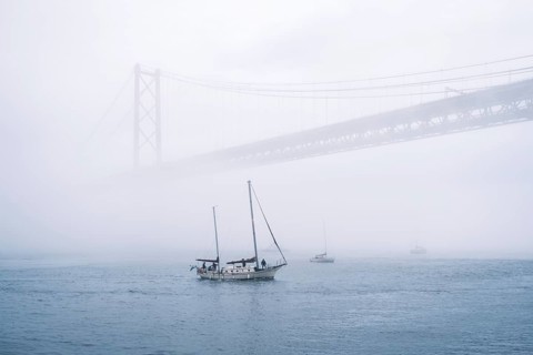 View of 25 de Abril Bridge famous tourist landmark of Lisbon connecting Lisboa and Almada in heavy fog mist wtih yacht boats passing under. Lisbon