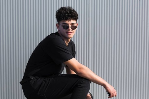 Young ethnic curly man sunglasses black clothes against grey wall