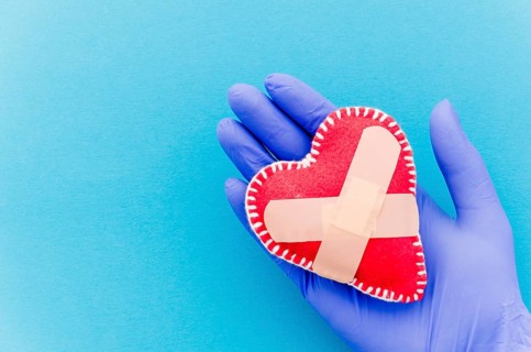 Close up hand wearing surgical gloves holding heart shaped stitched textile heart with crossed bandages blue backdrop