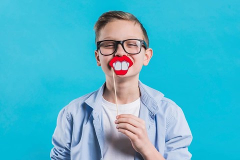 Boy wearing black eyeglasses holding smiling prop front his mouth against blue background
