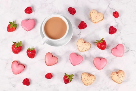 Pink and beige heart shaped French macaron sweets next to coffee cup and strawberry fruits