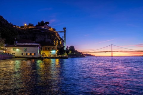 View of 25 de Abril Bridge famous tourist landmark of Lisbon over Tagus river and Boca do Vento Elevator and seaside restaurants with people in the evening. Lisbon