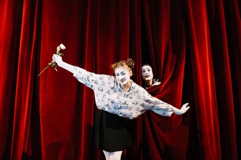 Female mime holding white rose performing stage with male mime peeking curtain
