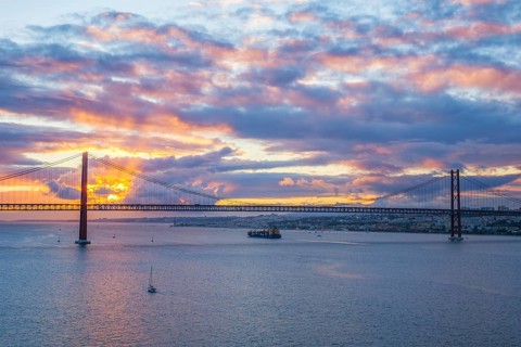 View of 25 de Abril Bridge famous tourist landmark of Lisbon over Tagus river with tourist yacht boats and cargo container ship on sunset. Lisbon