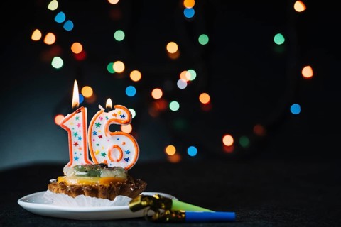 Number 16 birthday lighted candle slice tart with party horn blower against illuminated bokeh backdrop