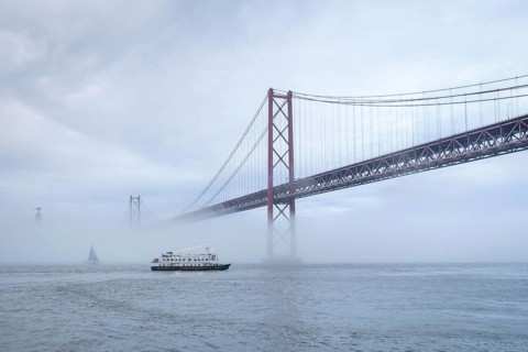 View of 25 de Abril Bridge famous tourist landmark of Lisbon connecting Lisboa and Almada in heavy fog mist wtih yacht boats passing under. Lisbon