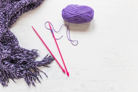 Knitted needles with purple yarn scarf white textured backdrop