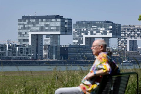 Senior couple sitting on a bench on the banks of the Rhine in front of the crane houses in Cologne's Rheinauhafen harbour on the other bank