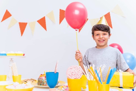 Smiling boy wearing party hat holding balloon gift standing variety food table