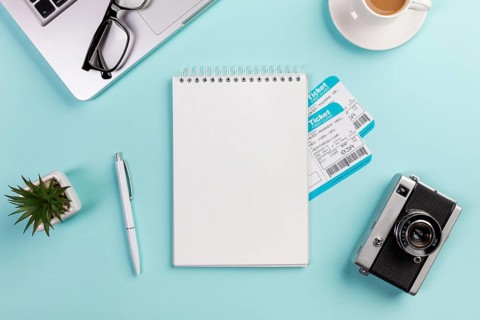 Blank spiral notepad with air tickets surrounded with laptop eyeglasses pen camera coffee cup blue desk