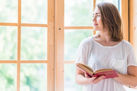 Pretty young woman standing front window holding book