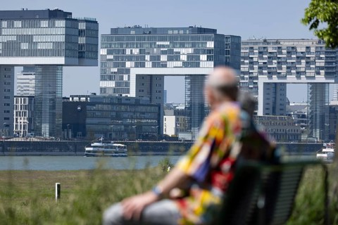 Senior couple sitting on a bench on the banks of the Rhine in front of the crane houses in Cologne's Rheinauhafen harbour on the other bank