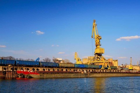 Colorful wide shot of river port crane loading open-top gondola cars on sunny day. Empty river drag boats or barges moored by pier. Empty cars ready for loading. Shipping raw materials