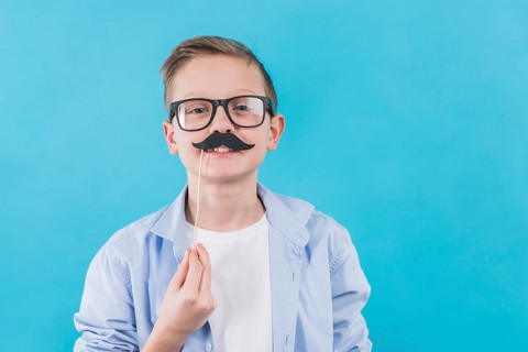 Boy wearing black eyeglasses holding black moustache prop front his upper lips